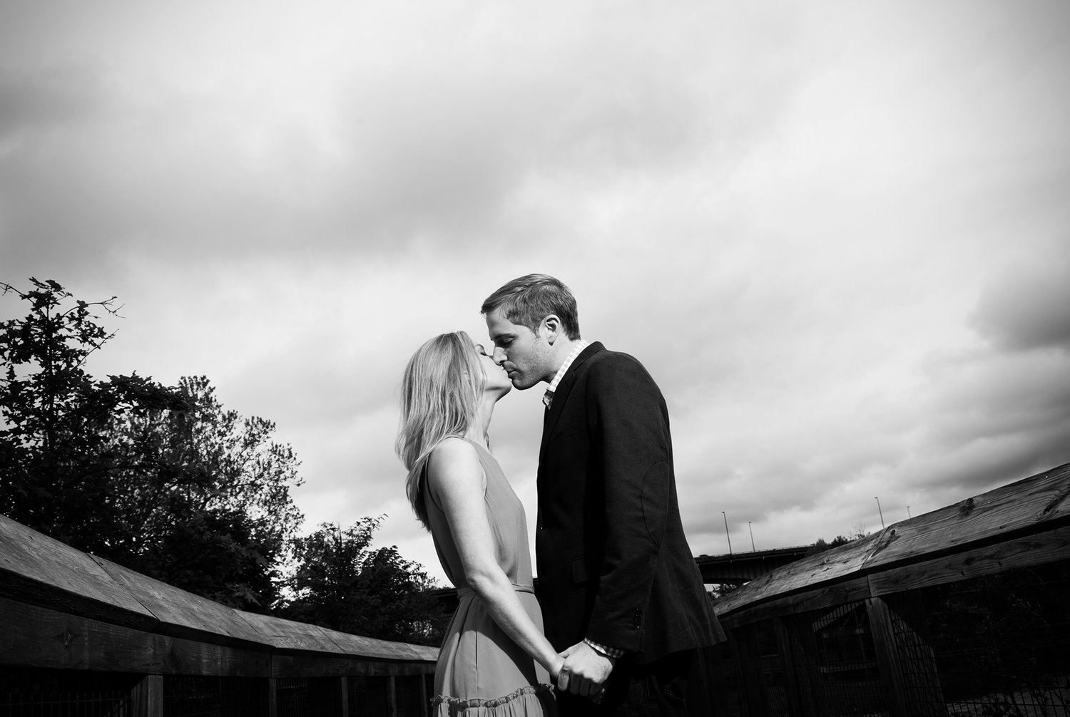 Black and white photograph of an engaged couple leaning in to kiss beside a wooden fence under a wide, cloudy sky