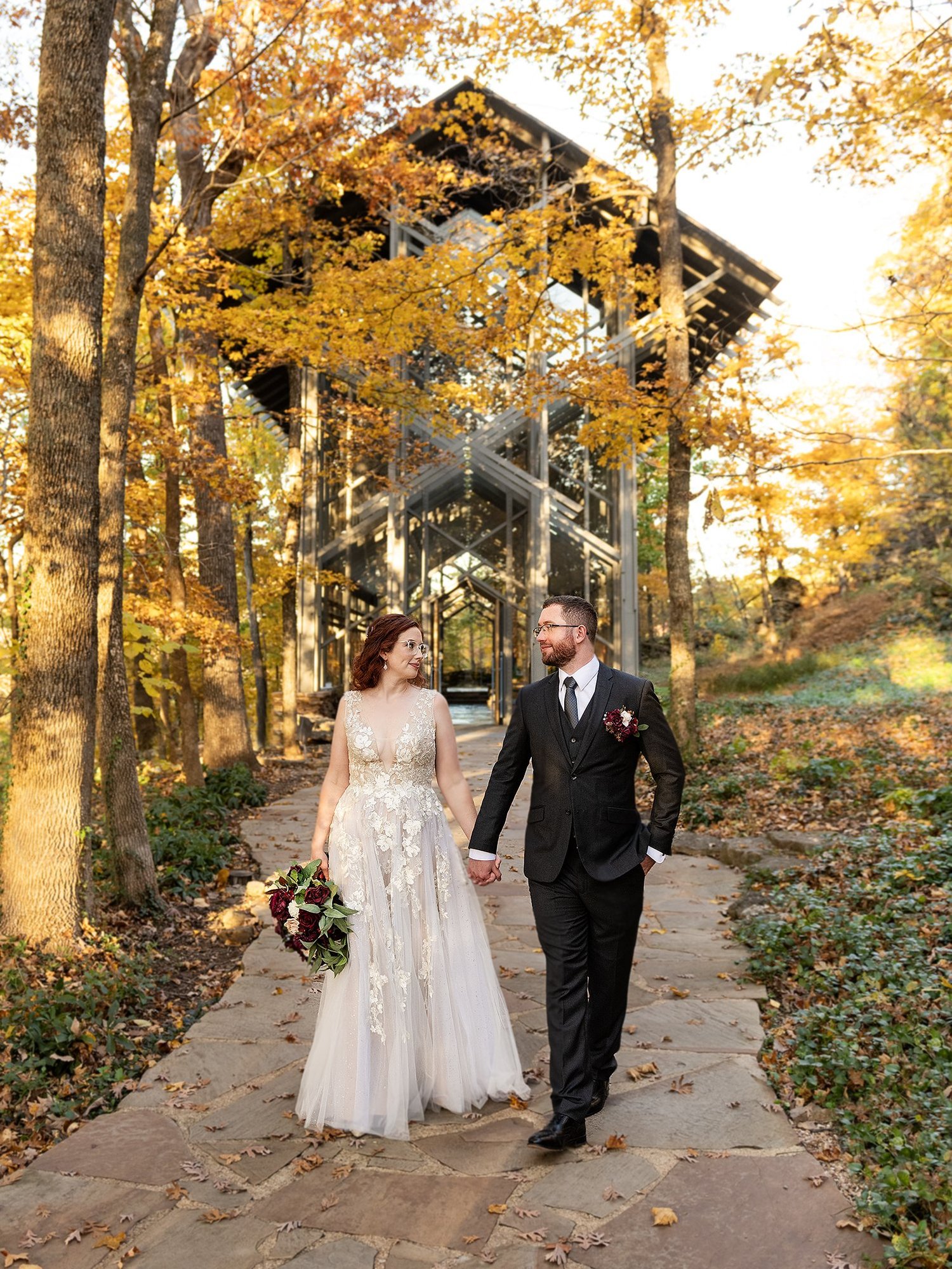 Bride and groom walking hand in hand down a flagstone path away from Thorncrown Chapel, autumn oaks in gold above them